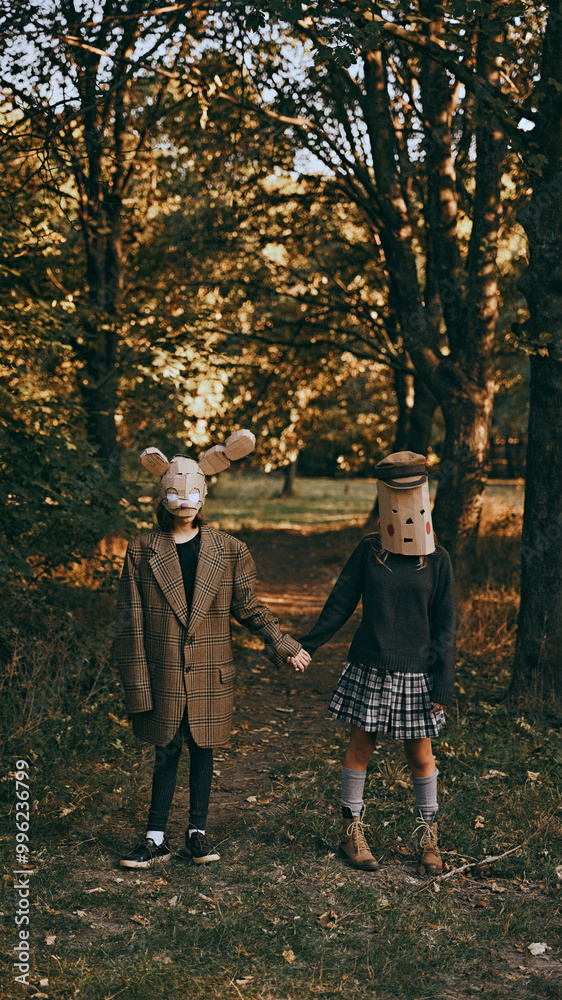 Two figures in oversized coats and strange, homemade masks stand in the autumn woods, creating creepy mystery of holiday. Concept of Halloween, spooky season, retro style. Two figures in oversized coats and strange, homemade masks stand in the autumn woods, creating creepy mystery of holiday. Concept of Halloween, spooky season, retro style.