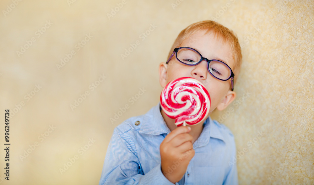 A young boy with glasses and squinting eyes is delightfully munching on a big red lollipop. A young boy with glasses and squinting eyes is delightfully munching on a big red lollipop.