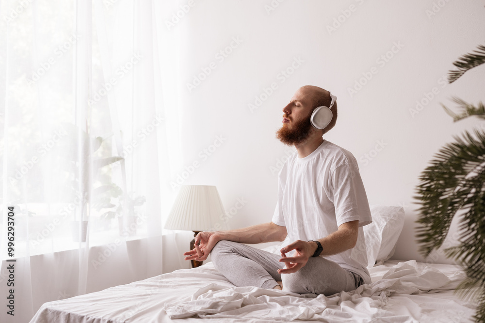 Young man meditating and doing yoga exercise using headphones sitting on bed at bedroom Young man meditating and doing yoga exercise using headphones sitting on bed at bedroom