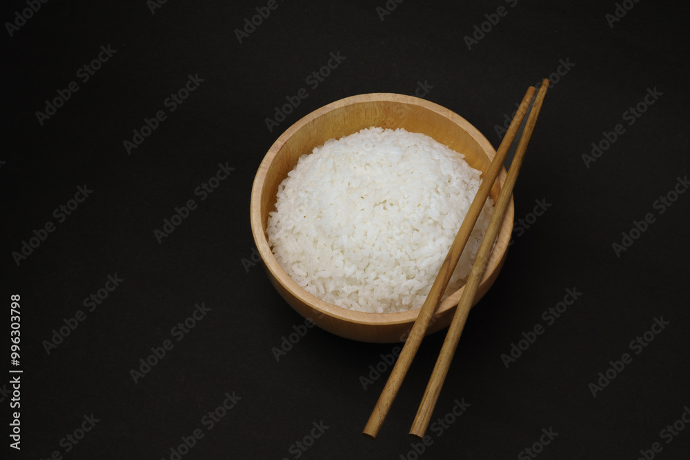 Wooden bowl filled with freshly steamed white rice with chopsticks on top, isolated on black background for minimalist food or culinary theme. Wooden bowl filled with freshly steamed white rice with chopsticks on top, isolated on black background for minimalist food or culinary theme.