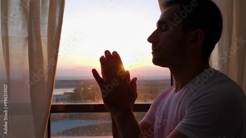 Man in prayer by a window during sunset, embracing a moment of reflection
