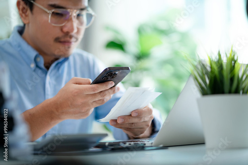 A man is sitting at a desk with a laptop and a cell phone. He is looking at the cell phone and he is checking something on it