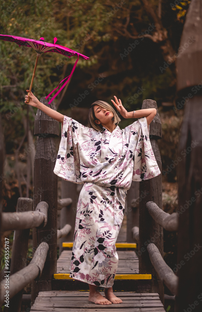 Fototapeta premium Tradition and beauty: japanese blonde woman in white silk kimono with flowers design and pink lace umbrella at wooden bridge in serene japanese park