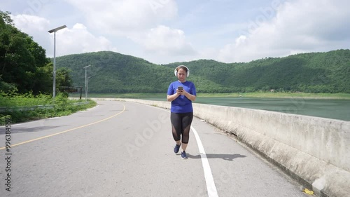 Wallpaper Mural A middle-aged, overweight Asian woman wearing headphones is walking for exercise on a road while using a smartphone, aiming for better health and weight loss, and relaxing during her workout Torontodigital.ca