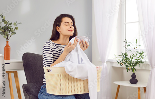 Young happy woman smelling fresh laundry aroma basket at home, clean clothes, white linen, housewife or maid spring cleaning with detergent, cleaner scent on fresh fabric, comfort sniff after washing 