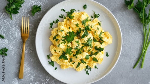 Scrambled eggs with green herbs and parsley on a white plate accompanied by a golden fork and knife displayed from a top view on a neutral grey table