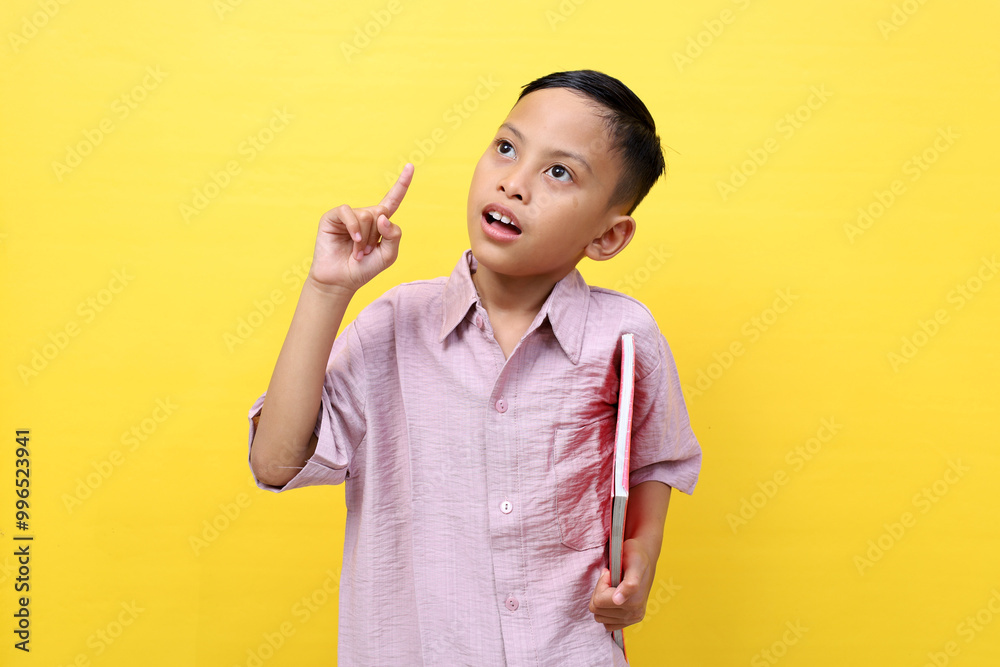 Happy asian boy standing while pointing finger and carrying a book. Isolated on yellow background Happy asian boy standing while pointing finger and carrying a book. Isolated on yellow background