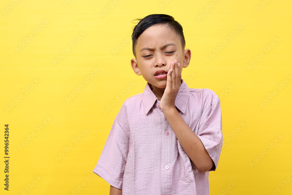 Asian kid suffering from toothache pain, holding his cheek, isolated on yellow background. Dental Health And Care Concept. Asian kid suffering from toothache pain, holding his cheek, isolated on yellow background. Dental Health And Care Concept.
