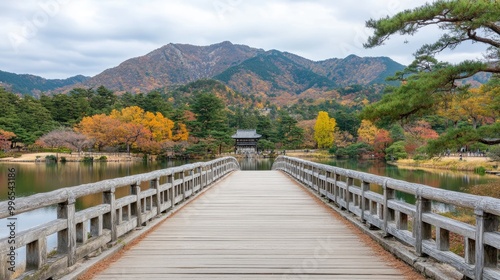 A wooden bridge welcomes visitors to a serene lake, framed by autumn foliage in red and orange, under clear blue skies and distant mountains, evoking tranquility