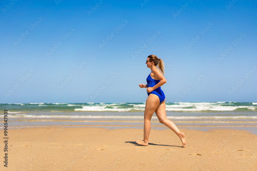 Beautiful middle-aged woman in one-piece blue swimsuit running on sandy beach in summertime. physical activity on beach. Side view Beautiful middle-aged woman in one-piece blue swimsuit running on sandy beach in summertime. physical activity on beach. Side view