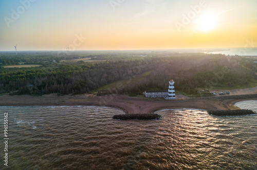 West Point lighthouse stands prominently on the coastline as the sun sets over the calm ocean waters, illuminating the sky with vibrant hues of orange and yellow, Prince Edward Island, Canada