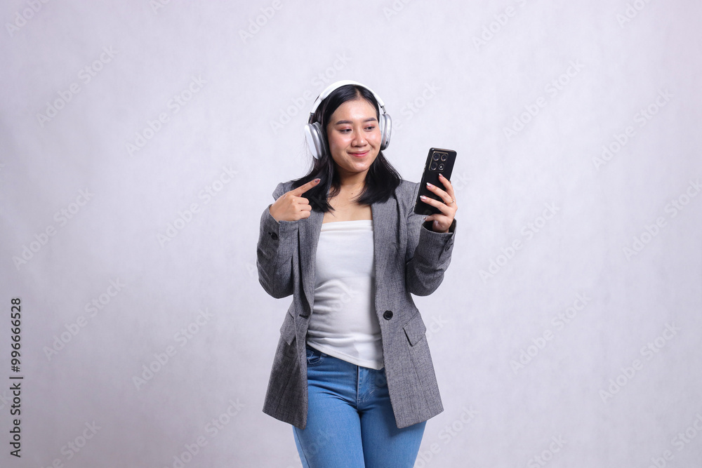 beautiful young office girl smile to gadget wearing headphone and holding mobile phone while pointing wearing suit isolated on white background. lifestyle concept, education, entertainment, technology beautiful young office girl smile to gadget wearing headphone and holding mobile phone while pointing wearing suit isolated on white background. lifestyle concept, education, entertainment, technology