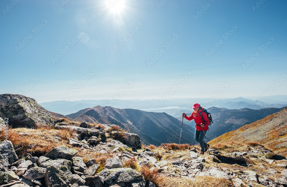 Backpacker woman in bright red jacket walking by mountain range using trekking poles with Liptov valley background, Western Tatras, Slovakia. Active people and European hiking tourism concept image.