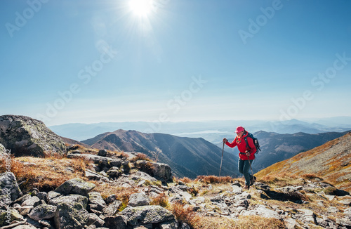 Backpacker woman in bright red jacket walking by mountain range using trekking poles with Liptov valley background, Western Tatras, Slovakia. Active people and European hiking tourism concept image.