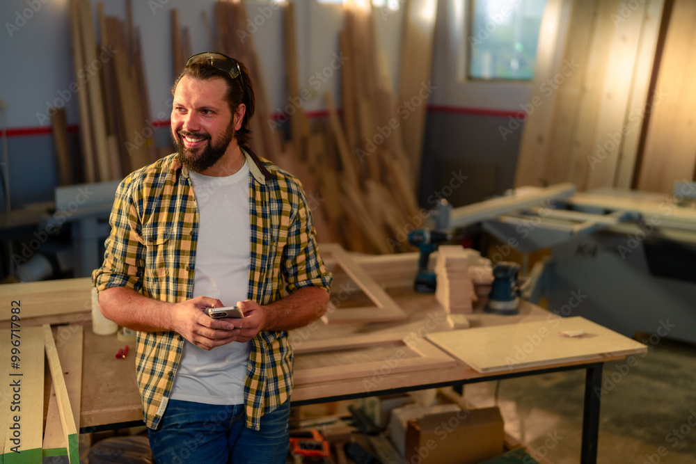 A carpenter in a workshop looking at his smartphone while leaning against a workbench. A carpenter in a workshop looking at his smartphone while leaning against a workbench.