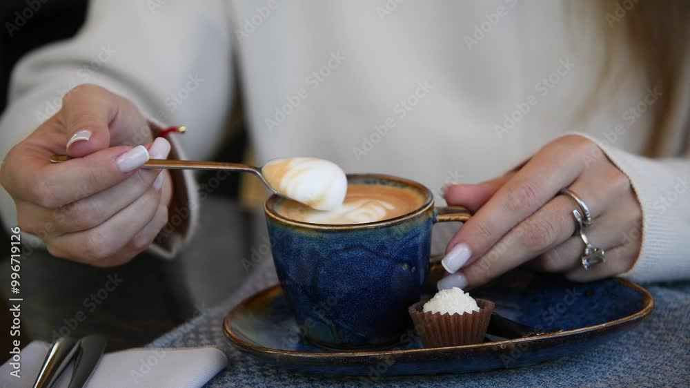 Close-up view of woman taking and mixing milk froth with teaspoon in blue porcelain cup full of cappuccino or latte coffee drink in restautant or cafe. Soft focus. Food and elegance theme. Close-up view of woman taking and mixing milk froth with teaspoon in blue porcelain cup full of cappuccino or latte coffee drink in restautant or cafe. Soft focus. Food and elegance theme.