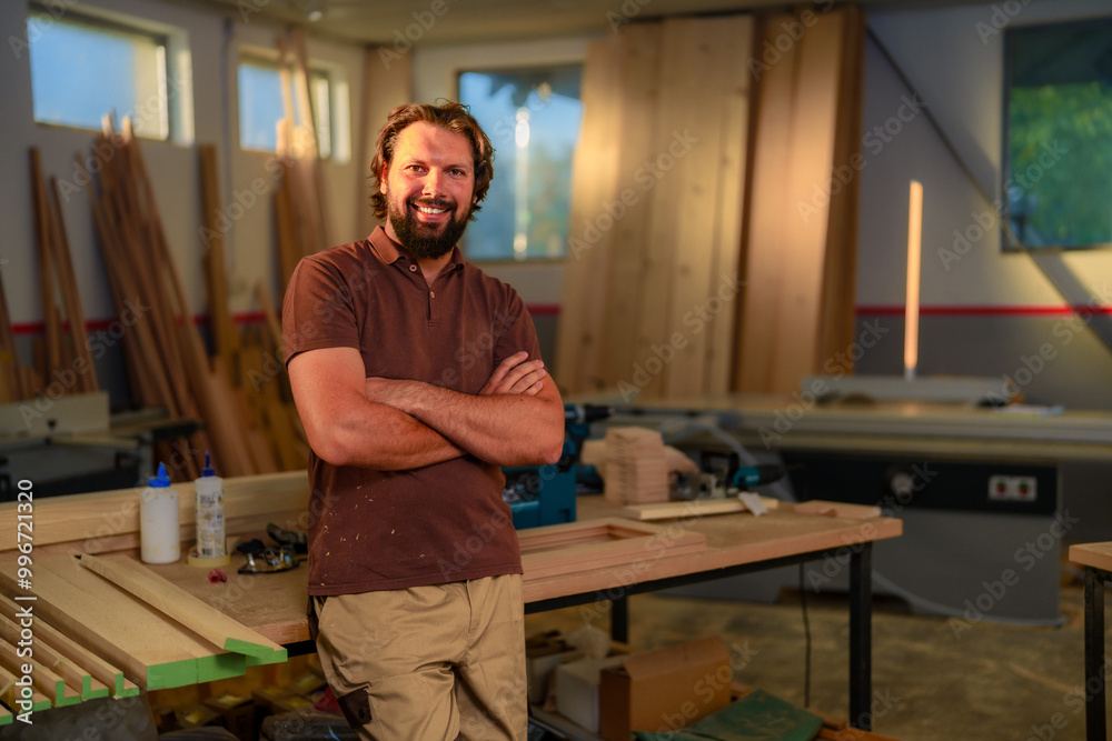 A portrait of smiling carpenter who stands with crossed hands in a woodworking shop, dressed in working suit, surrounded by stacks of lumber and leaned on working table. A portrait of smiling carpenter who stands with crossed hands in a woodworking shop, dressed in working suit, surrounded by stacks of lumber and leaned on working table.