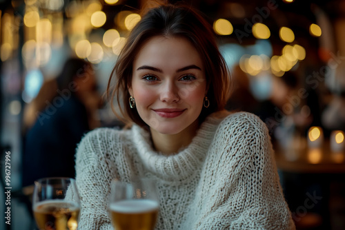 Cute woman in her early twenties, wearing a white knit sweater and earrings, sitting at a table with a glass of wine. photo