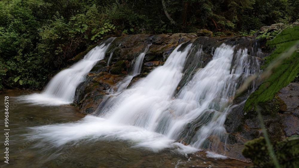 This small waterfall gently flows over rocks in a lush forest setting, surrounded by vibrant green foliage. The smooth, flowing water creates a calming and peaceful atmosphere in nature. This small waterfall gently flows over rocks in a lush forest setting, surrounded by vibrant green foliage. The smooth, flowing water creates a calming and peaceful atmosphere in nature.