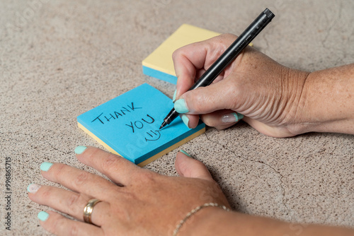 A close-up of a person writing Thank you on a blue sticky note with a black pen. The person's hands and a stack of sticky notes are visible on a textured surface.