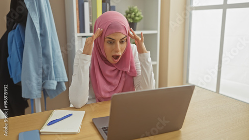 Shocked muslim woman in hijab at home office staring at laptop screen, expressing surprise.