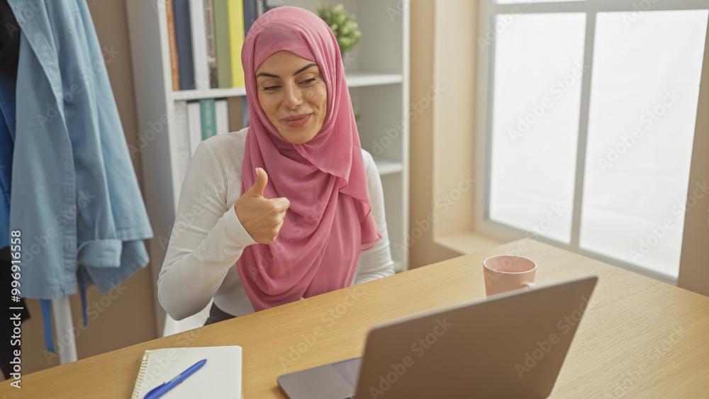 A smiling woman wearing hijab gesturing thumbs-up at a desk with a laptop, in a bright home office setting. A smiling woman wearing hijab gesturing thumbs-up at a desk with a laptop, in a bright home office setting.