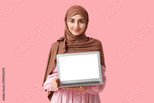 Beautiful Muslim woman with modern laptop on pink background