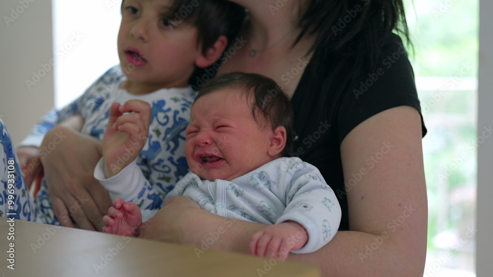 Mother holding her baby and young child, comforting a crying baby. family is sitting at a table, showcasing the challenges and love in parenthood. Children are in cozy pajamas Mother holding her baby and young child, comforting a crying baby. family is sitting at a table, showcasing the challenges and love in parenthood. Children are in cozy pajamas