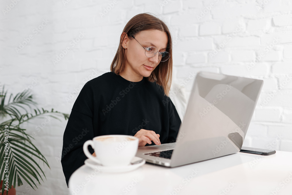 Smiling young woman freelancer brows internet on laptop while drinks coffee at workplace. Female entrepreneur, businesswoman or manager distance works online watches webinars, student studies remote Smiling young woman freelancer brows internet on laptop while drinks coffee at workplace. Female entrepreneur, businesswoman or manager distance works online watches webinars, student studies remote