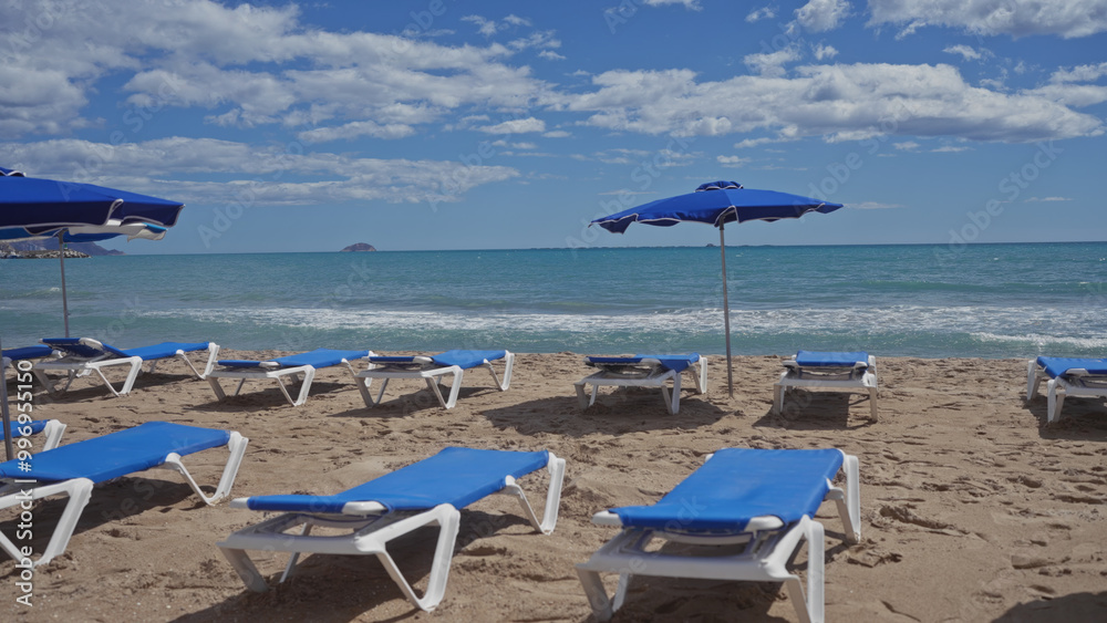 Blue sunbeds and umbrellas on a sandy beach overlooking the calm sea under a cloudy sky, depicting a typical summer vacation scene. Blue sunbeds and umbrellas on a sandy beach overlooking the calm sea under a cloudy sky, depicting a typical summer vacation scene.