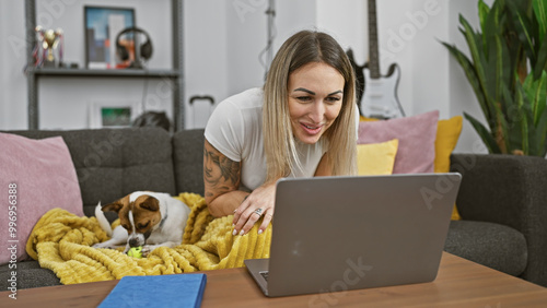 A young woman with a tattoo smiles as she works on her laptop at home, with her dog playing nearby. photo