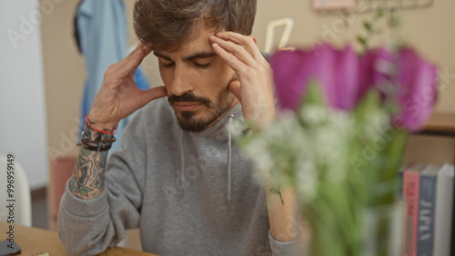 A young bearded hispanic man in a grey sweater feeling stressed at home seated with his eyes closed and hands on his forehead.