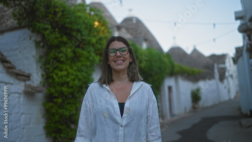 A beautiful young hispanic woman smiles on a quaint street in alberobello, puglia, italy, surrounded by traditional trulli houses and lush greenery.