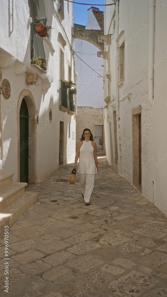 Naklejka premium A beautiful young hispanic woman walks through the charming old streets of locorotondo, puglia, italy, surrounded by white buildings and european architecture.