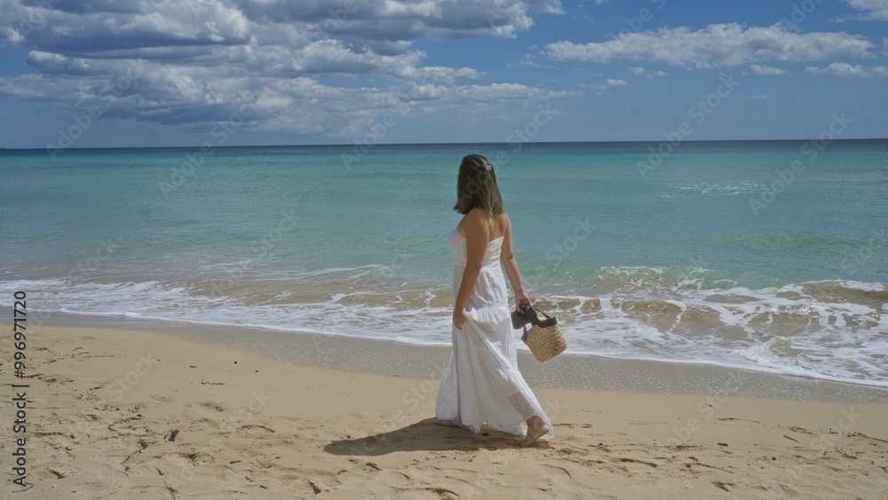 A young, beautiful, hispanic woman in a white dress walks along the beach in pescoluse, puglia, italy, carrying a straw bag with clear blue skies and calm sea waters in the background. A young, beautiful, hispanic woman in a white dress walks along the beach in pescoluse, puglia, italy, carrying a straw bag with clear blue skies and calm sea waters in the background.