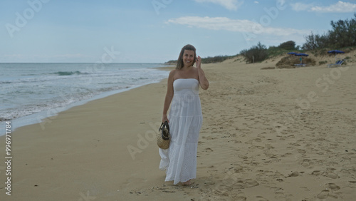 A beautiful young hispanic woman walks along the serene beach of pescoluse in puglia, italy, dressed in a flowing white sundress and carrying a straw bag. photo