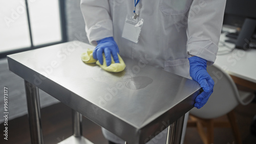 A woman wearing blue gloves cleans a stainless steel table in a clinic or hospital room with a yellow cloth, indicating a sanitary indoor workplace environment.
