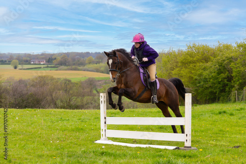 Up and over, young woman and her pretty bay horse pony enjoy jumping home made jumps in the English countryside on a summers day. photo