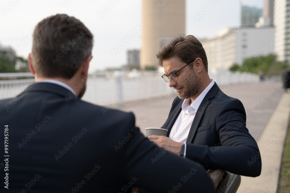 Business meeting of friends outdoors. Two men in suits are sitting on a bench near a city building with a laptop and talking. Partnership and corporate relations. Smiling partners discussing Business meeting of friends outdoors. Two men in suits are sitting on a bench near a city building with a laptop and talking. Partnership and corporate relations. Smiling partners discussing