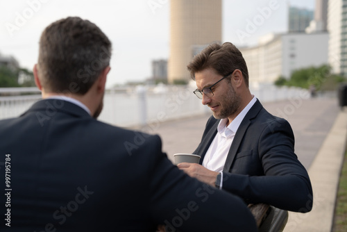 Business meeting of friends outdoors. Two men in suits are sitting on a bench near a city building with a laptop and talking. Partnership and corporate relations. Smiling partners discussing