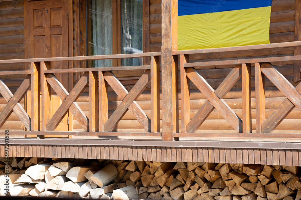 Ukrainian blue and yellow flag hang on wooden terrace, fragment of house with wood fence. Eco house made from natural material Ukrainian blue and yellow flag hang on wooden terrace, fragment of house with wood fence. Eco house made from natural material