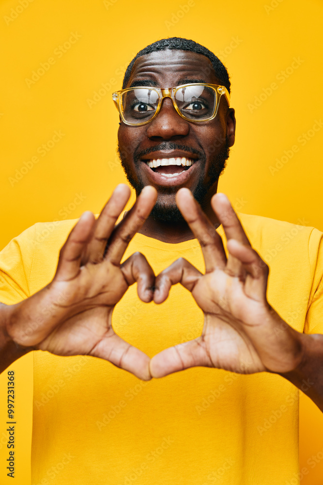 Happy young man making heart shape with hands, wearing yellow t shirt and glasses, bright yellow background, cheerful emotion, positive vibes, joyful expression, joyful concept Happy young man making heart shape with hands, wearing yellow t shirt and glasses, bright yellow background, cheerful emotion, positive vibes, joyful expression, joyful concept