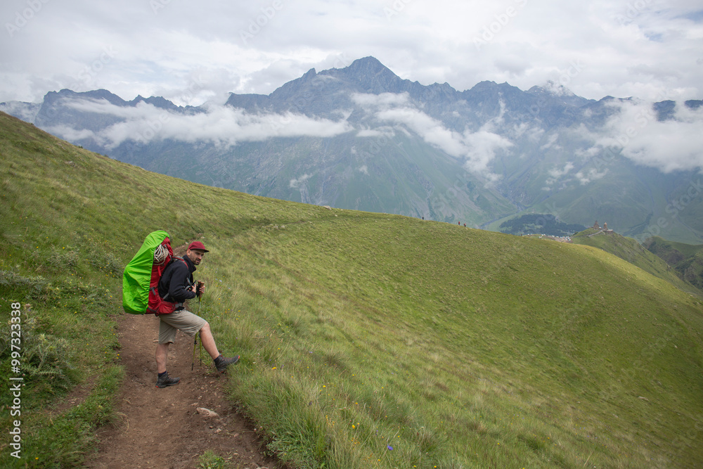 A hiker enjoys a peaceful moment on a scenic trail, surrounded by green hills and towering mountains. A hiker enjoys a peaceful moment on a scenic trail, surrounded by green hills and towering mountains.