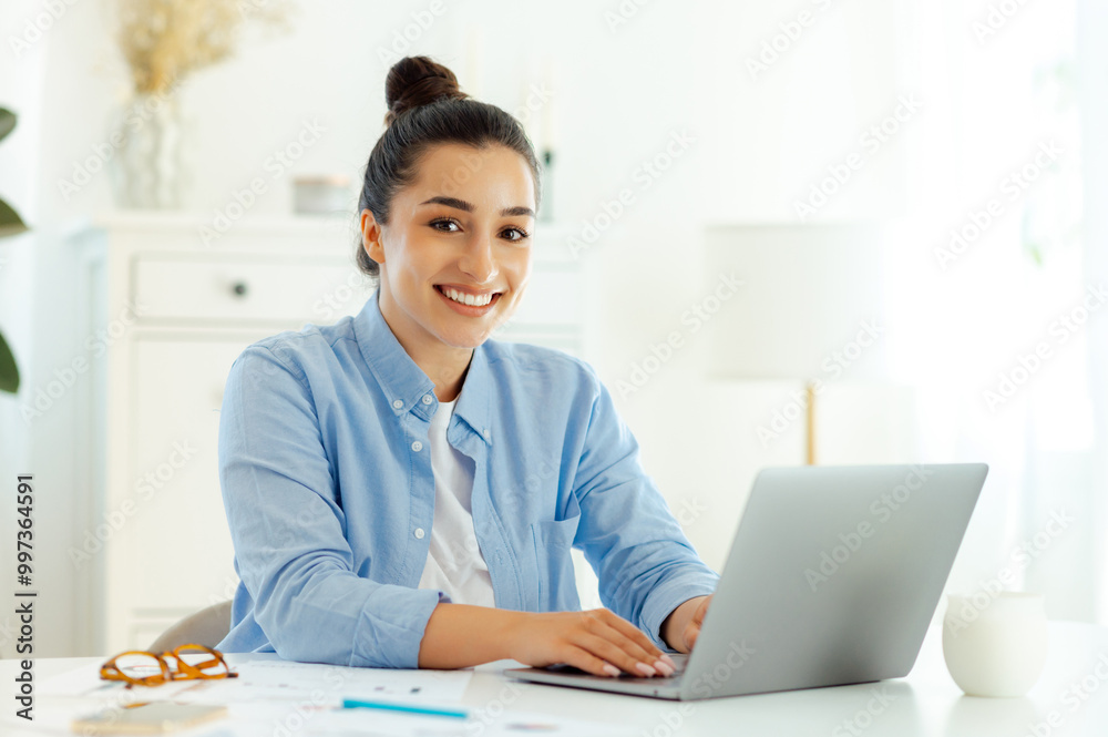 Confident attractive successful caucasian young brunette woman in a blue shirt, company worker, business lady, sitting at her workplace with laptop, looking at the camera, smiling friendly Confident attractive successful caucasian young brunette woman in a blue shirt, company worker, business lady, sitting at her workplace with laptop, looking at the camera, smiling friendly