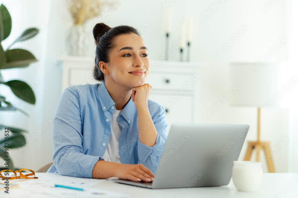 Dreamy happy caucasian young woman in a blue shirt, company worker, sitting at her workplace with laptop, thoughtfully looking away and smiling, thinking about project profit, dreaming about weekend Dreamy happy caucasian young woman in a blue shirt, company worker, sitting at her workplace with laptop, thoughtfully looking away and smiling, thinking about project profit, dreaming about weekend