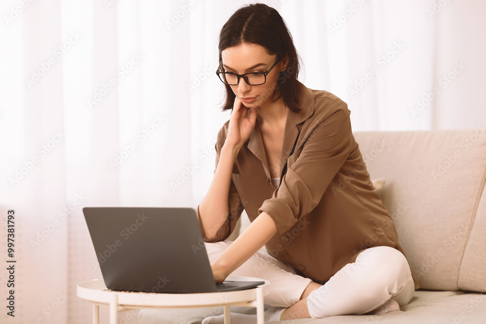 Freelance. Casual beautiful young woman in eyewear working on a laptop sitting on the couch at home, copyspace Freelance. Casual beautiful young woman in eyewear working on a laptop sitting on the couch at home, copyspace