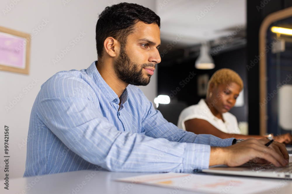 Working on laptop, african american man focusing on task while woman writing in background Working on laptop, african american man focusing on task while woman writing in background
