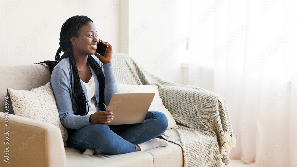 Recruitment concept. Unemployed african american woman talking on cellphone and holding laptop, scheduling job interview while sitting on sofa at home Recruitment concept. Unemployed african american woman talking on cellphone and holding laptop, scheduling job interview while sitting on sofa at home