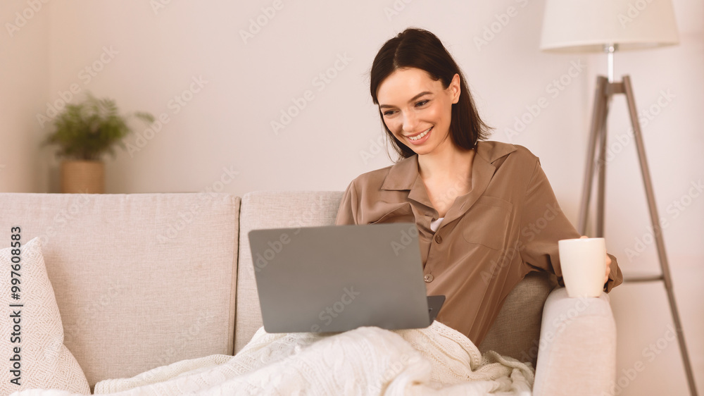Good Morning. Smiling student sitting on couch drinking coffee at home and using laptop, watching video, copyspace Good Morning. Smiling student sitting on couch drinking coffee at home and using laptop, watching video, copyspace