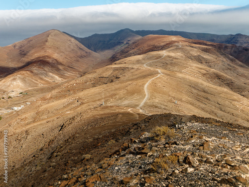 The trail to the summit of Pico de la Zarza, the highest mountain on Fuerteventura, winds through arid, rocky terrain. The undulating landscape reveals distant hills, with a cloud-topped horizon in th